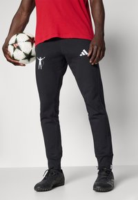 Juodi sportiniai kelnių su kontrastinga balta Adidas logotipo ir grafika. Fiksuota dizaino. Dėvimi su juodais futbolo batais, laikant raštuotą futbolo kamuolį.