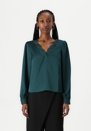 Tyrkysfarget bluse med lange ermer og blonder langs V-halsen og mansjettene. Laget av et glatt stoff, har en løs passform.
