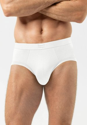 Mannelijk bovenlichaam en bovenbenen gekleed in effen witte slips met gekruiste armen over de borst tegen een effen achtergrond.