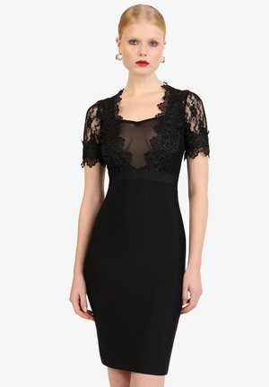 Frau trägt ein figurbetontes schwarzes Kleid mit transparenten Spitzen-Ärmeln und floraler Spitzenborte am Ausschnitt und oberen Brustbereich.