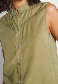 Blusa sin mangas color verde oliva con un cuello alto, que presenta costuras decorativas y botones plateados en la parte delantera. Textura suave.