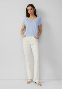 Jasnoniebieska koszulka z dekoltem w serek i krótkimi rękawami, zestawiona z białymi spodniami o szerokich nogawkach. Model stoi na neutralnym tle, z rękami w kieszeniach.