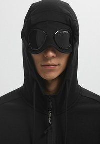 Felpa nera con colletto alto e chiusura con zip, abbinata a occhiali da moto rotondi e scuri con un logo. Il tessuto sembra morbido e testurizzato.