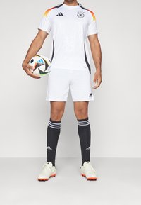 Manlig fotbollsspelare i vit Tysklandströja och shorts, svarta strumpor, vita fotbollsskor, som håller en flerfärgad fotboll vid sin sida.