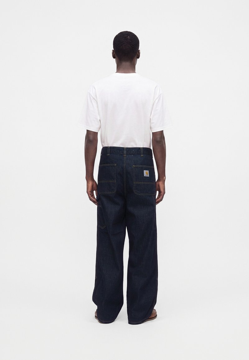 Man wearing a white short-sleeve shirt and dark blue loose-fitting jeans standing facing away on a plain white background.