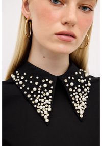 Sort bluse med en struktureret krave prydet med perler i forskellige størrelser og klare krystaller, der skaber en tekstureret, dekorativ effekt.
