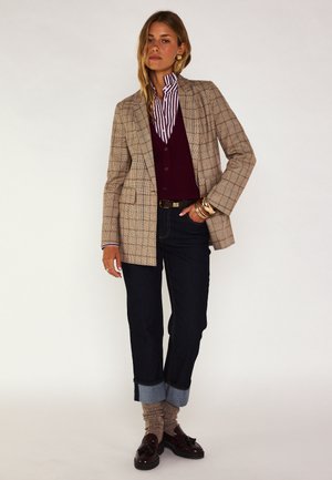Femme portant un blazer beige à carreaux, une chemise rayée, un gilet bordeaux, un jean foncé avec revers, des chaussettes beiges et des mocassins foncés, debout devant un fond uni.