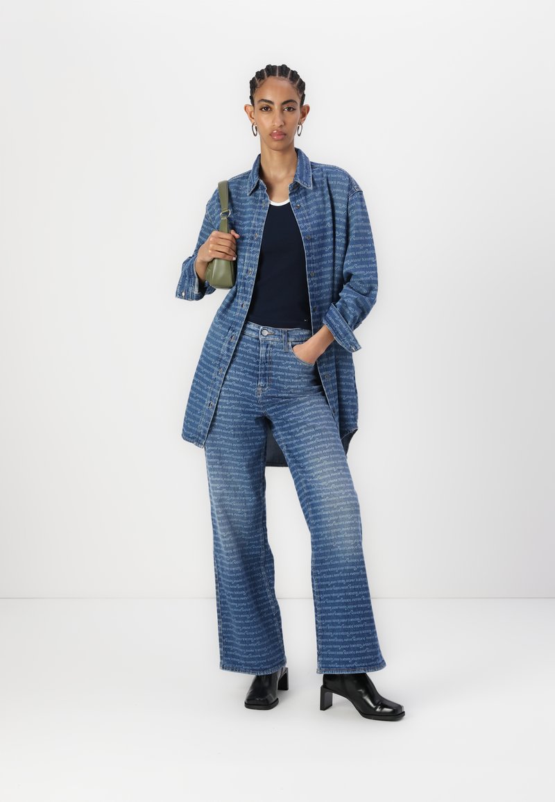 Denimhemd und -hose mit hellblauem und weißem Textmuster; kombiniert mit einem dunklen Oberteil und einer grünen Tasche; schwarze Absatzstiefel; lockere Passform.