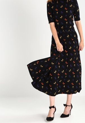 Mujer con vestido midi negro con patrones de lazos multicolores, sosteniendo la falda, combinado con tacones altos negros con correa en el tobillo.