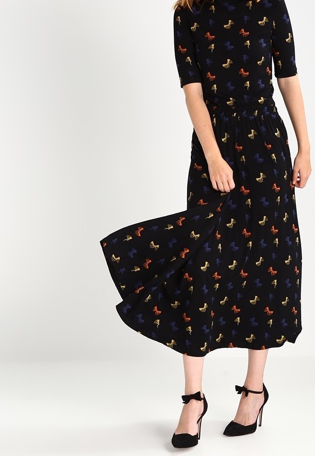 Femme portant une robe midi noire avec des motifs de nœuds multicolores, tenant la jupe, associée à des talons hauts noirs à bride cheville.