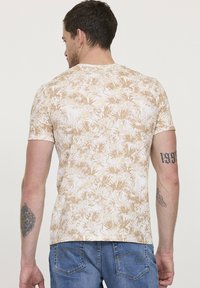 T-shirt à manches courtes en blanc avec un motif de feuilles tropicales beige. Tissu doux, design ajusté et col rond, avec des tatouages visibles sur le bras.