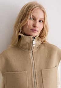 Beige fleecevest met hoge kraag en zilveren rits. Voorzien van twee zakken aan de voorkant. Zachte textuur en minimalistisch ontwerp.