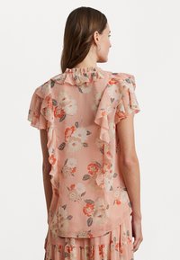 Blomstrete bluse i lys rosa med volanger på skuldrene. Har trykte blomster i oransje og krem mot et strukturert stoff.