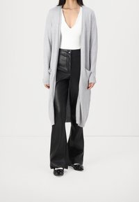 Frau trägt einen hellgrauen langen Cardigan, ein weißes V-Ausschnitt-Oberteil, schwarze weit geschnittene Lederhosen und schwarze spitze Schuhe mit Lochdetails.