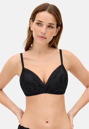 Mujer de piel clara y cabello castaño que lleva un sujetador de encaje negro, de pie con las manos en las caderas sobre un fondo neutro.