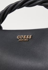 Svart handväska i konstläder med en vriden handtag, prydd med en guldig logotyp som lyder "GUESS EST 1981" präglad på framsidan.