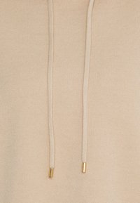 Beige Pullover mit strukturiertem Ripp-Design, ausgestattet mit einer Kapuze und Kordeln mit goldenen Metallspitzen. Glattes Material mit subtiler Schimmeroberfläche.