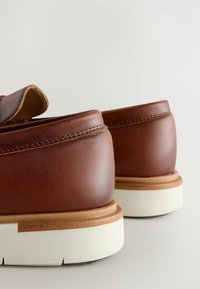 Chaussures en cuir marron avec une texture lisse, des bords arrondis, et des semelles en caoutchouc blanches présentant un accent marron autour de la semelle intermédiaire.
