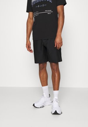 Mężczyzna w czarnej koszulce z nadrukiem, czarnych krótkich spodenkach za kolano, białych skarpetkach do kostek oraz białych sportowych butach stojący na gładkiej podłodze.