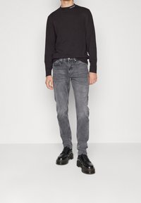 Chemise noire à manches longues avec logo, jean gris légèrement délavé, et bottes noires à lacets avec semelle épaisse. Style simple et moderne avec des lignes épurées.