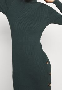 Ribbed, tummanvihreä poolomekko, jossa on sivunappiyksityiskohta. Kiinteä siluetti, pitkät hihat ja pehmeä tekstuuri.