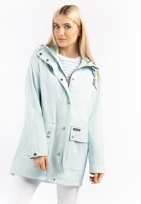 Fiatal nő világoskék esőkabátot visel, ezüst gombokkal és elülső zsebekkel, fehér háttér előtt áll.