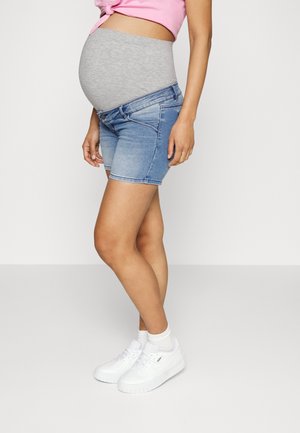 Mujer embarazada que lleva pantalones cortos premamá grises debajo de pantalones cortos vaqueros, top rosa anudado, calcetines blancos y zapatillas blancas, de pie de lado.