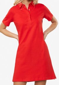 Robe polo rouge à manches courtes, col et trois boutons. Comprend un corsage ajusté et une jupe évasée, fabriquée dans un tissu lisse.