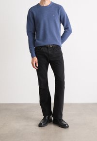 Maglione blu a collo rotondo con un piccolo logo, abbinato a jeans neri a gamba dritta e scarpe nere stringate, su uno sfondo neutro.