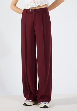 Femme portant un pantalon taille haute à jambes larges bordeaux, associé à des baskets blanches et violettes et un top blanc court.