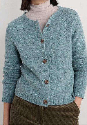 Femme portant un cardigan en maille bleu sarcelle avec des boutons marron sur un col roulé beige et un pantalon en velours côtelé marron, mains dans les poches.