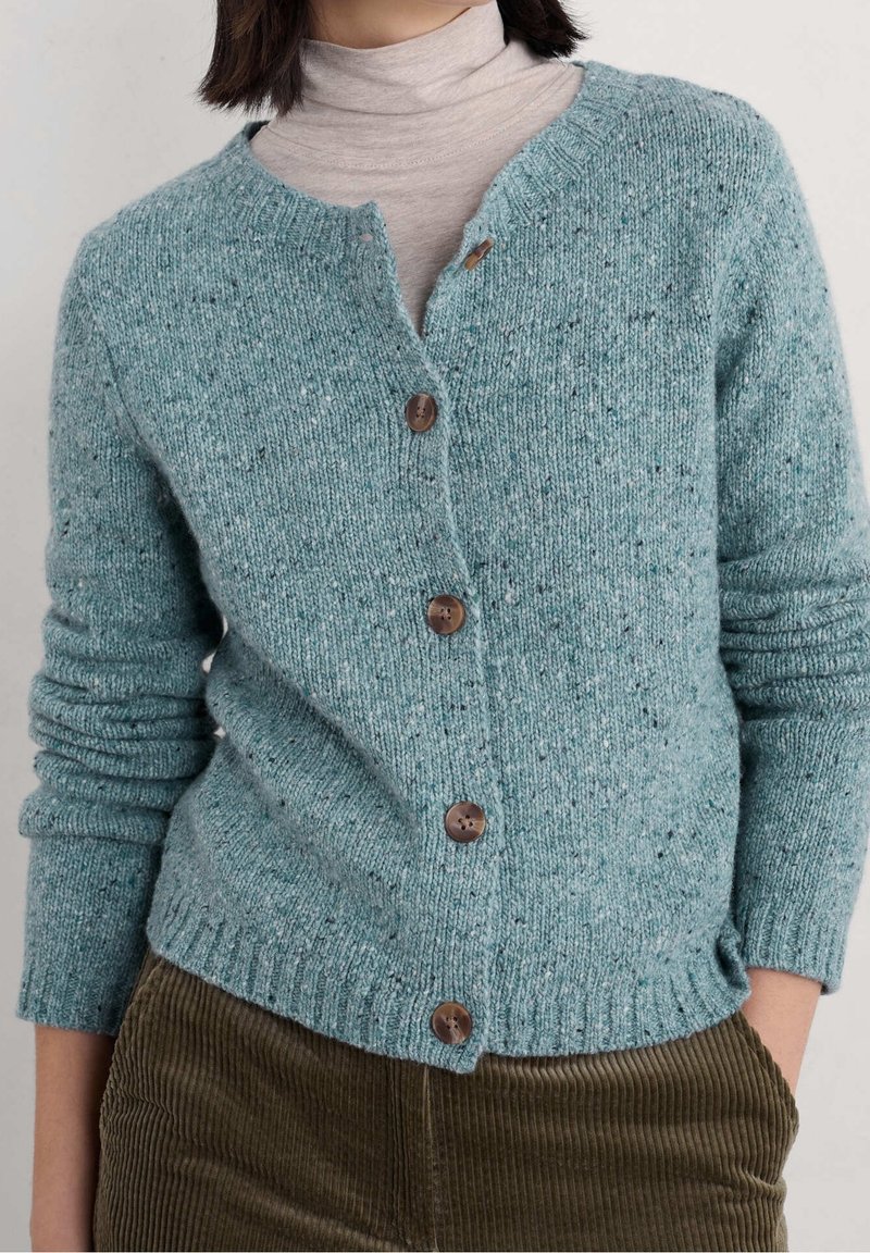 Femme portant un cardigan en maille bleu sarcelle avec des boutons marron sur un col roulé beige et un pantalon en velours côtelé marron, mains dans les poches.