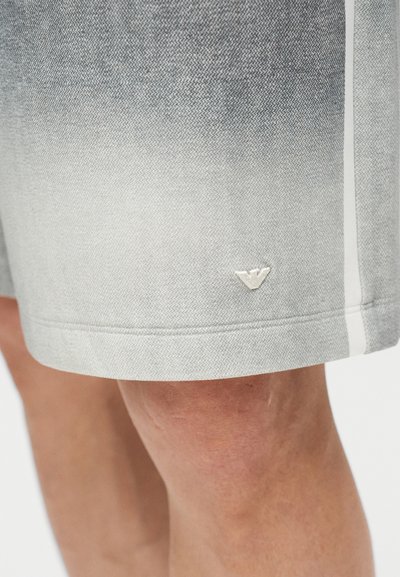 Γκρίζες σορτς με λευκή κάθετη ρίγα στο πλάι, που φοριούνται από ένα άτομο, με ένα μικρό λευκό λογότυπο κοντά στο τελείωμα.