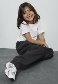 Enfant assis en tailleur sur une surface grise, portant un t-shirt en coton blanc, un pantalon noir et des baskets blanches avec des accents gris.
