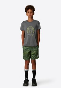 Camiseta de algodón gris con gráfico geométrico amarillo, combinada con pantalones cortos verdes con estampado. Zapatillas negras y calcetines hasta la rodilla completan el atuendo.