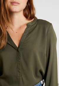 Blouse vert olive avec un col en V, une fermeture à boutons et des manches longues. Le tissu a une texture lisse avec un éclat subtil.