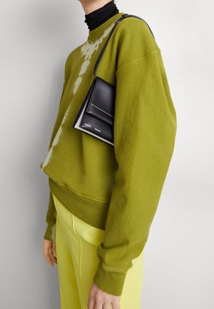Grön sweatshirt med batikmönster, tillsammans med gula byxor och en liten svart läderhandväska med vita sömmar.