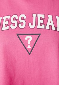 Světlý růžový tričko s textem „GUESS JEANS“ v bílé barvě, s černým trojúhelníkem obsahujícím bílý otazník a obrácený trojúhelník.