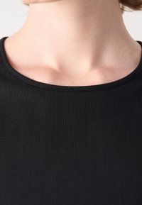 Gros plan sur le cou et le haut de la poitrine d'une personne portant un t-shirt noir à col rond sur un fond uni.