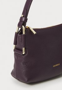 Borsa a tracolla in pelle viola con finitura testurizzata, dettagli in ottone dorato, chiusura zip e una tracolla corta e regolabile.