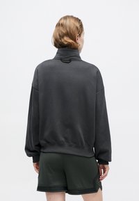 Grijze sweatshirt met hoge kraag, geribbelde onderrand en verlaagde schouders, gecombineerd met donkergroene textuuroveralls. Eenvoudig, modern ontwerp.