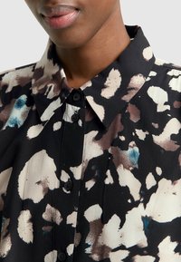 Blusa negra con cuello, que presenta un patrón irregular de manchas en tonos de crema, marrón y azul, y cinco botones negros visibles.