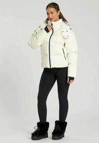 Witte pufferjas met een hoge kraag, ritssluiting en zwarte accenten, gedragen over zwarte leggings en zwart met bont afgewerkte laarzen.