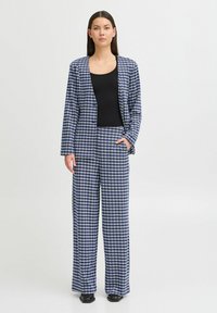 Navy- und weiße karierte Blazerjacke mit passenden weit geschnittenen Hosen, die einen lässigen Schnitt und eine subtile Textur aufweisen. Darunter ein schwarzes, tailliertes Oberteil.