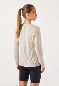 Lichtbeige performance top met lange mouwen, gestructureerde stof, zijnaad en reflecterende accenten, gecombineerd met zwarte, nauwsluitende shorts.