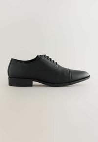 Zapato de vestir de cuero negro con superficie texturizada, puntera afilada y cierre de cordones de cinco ojetes. Presenta un tacón bajo y rectangular.