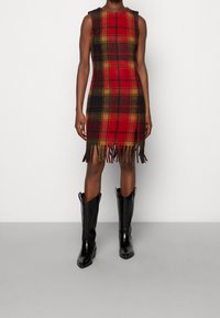 Robe à carreaux rouge et noir avec un ourlet à franges, sans manches, fabriquée en tissu en mélange de laine, associée à des bottes noires montant au genou.