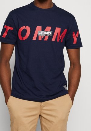 Mężczyzna w granatowej koszulce z czerwonymi literami "TOMMY" i białą etykietą "JEANS" na piersi, w połączeniu z beżowymi spodniami, ręce w kieszeniach.