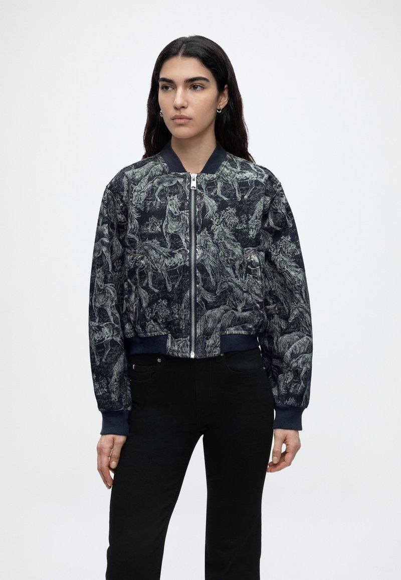 Γυναίκα με μαύρο bomber μπουφάν με γκρι σχέδιο αλόγου, κλεισμένο με φερμουάρ, συνδυασμένο με μαύρο παντελόνι, που στέκεται μπροστά σε απλό φόντο.