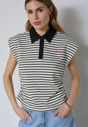 Žena nosiaca čierno-biele pruhované bezrukávové polo s červeným vyšívaným nápisom „Coco“, svetlomodré rifle, zlaté náušnice kruhy a náramok.